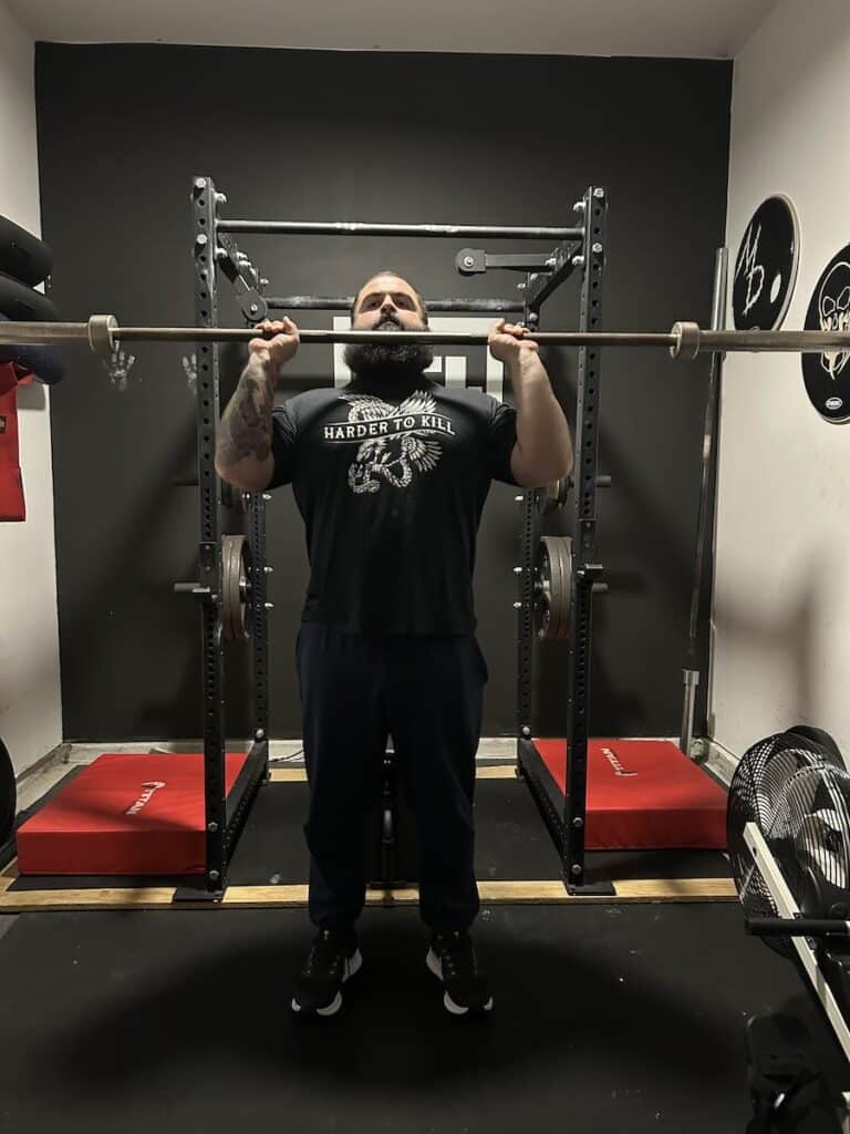 Strength coach shows how to do shoulder press with a barbell.