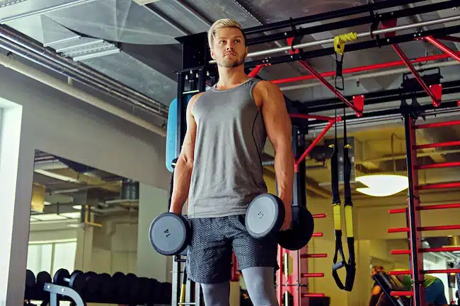 Blond sporty male doing squats with dumbbells in a gym club.