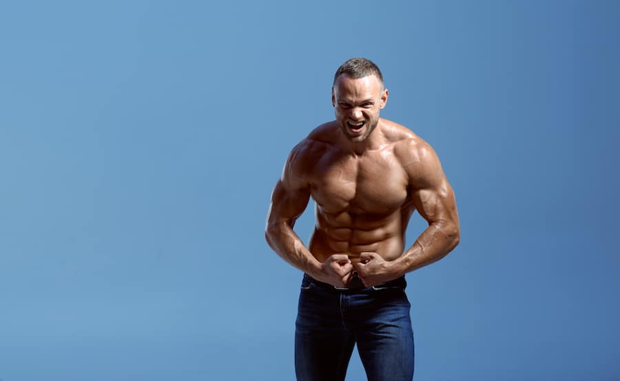 Male athlete with muscular body shows his biceps in studio, blue background. One man with athletic build, shirtless sportsman in jeans pants, active healthy lifestyle