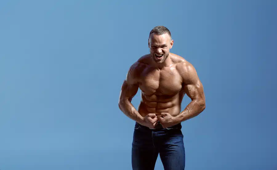 Male athlete with muscular body shows his biceps in studio, blue background. One man with athletic build, shirtless sportsman in jeans pants, active healthy lifestyle