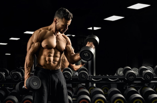 Front view of shirtless bodybuilder training biceps with weights near stand with dumbbells. Close up of muscular sportsman with perfect body posing in gym in dark atmosphere. Concept of bodybuilding.