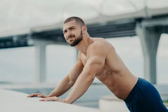 Side view of athletic motivated purposeful bearded sportive bare chested man performs push ups exercise, makes efforts leans on bridge hence looks into distance, has muscular body, works out outside.