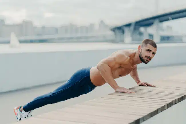Full length shot of sporty muscular man in sportswear does push up exercise has concentrated serious expression poses outdoor. Self determined bearded sportsman planking for having strong arms