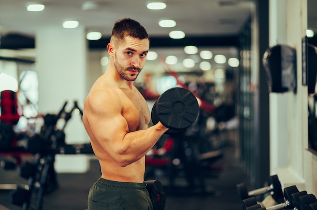A strong shirtless muscular sportsman is flexing muscles and lifting a dumbbell while making eye contact at the gym.
